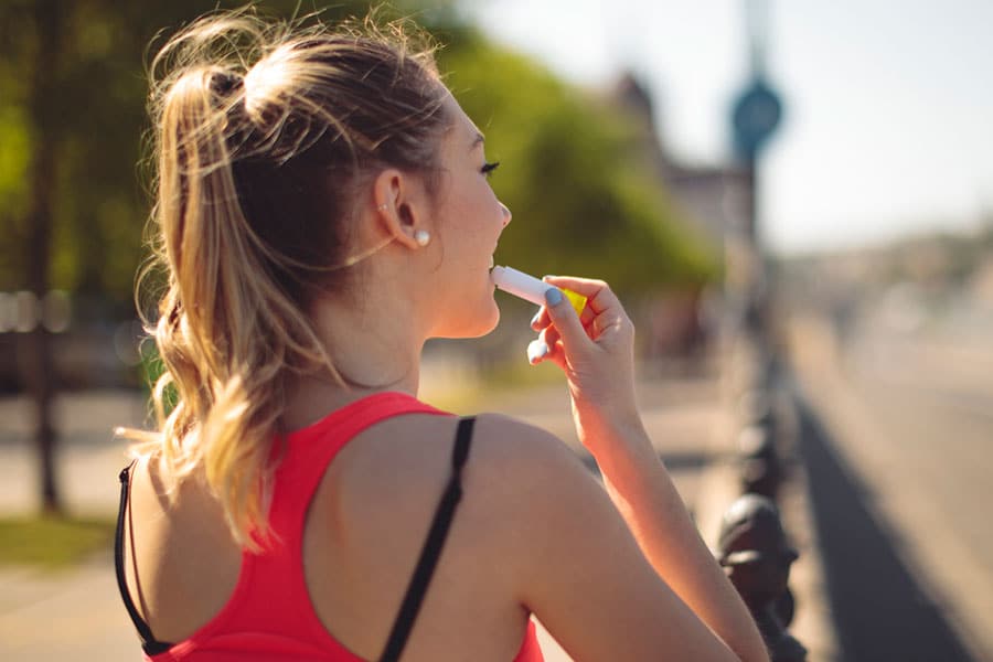 A woman with blonde hair in a ponytail applies lip balm—an essential part of many cold sore remedies—while standing outdoors in a bright, sunny setting, wearing a pink tank top. The background features blurred trees and a street.