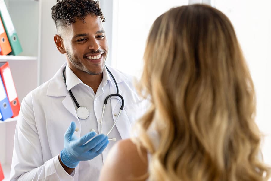 A smiling male doctor wearing a white coat, stethoscope, and blue gloves talks to a woman with long, wavy hair in a medical office, emphasizing the importance of your health. Colorful folders are visible on a shelf in the background.