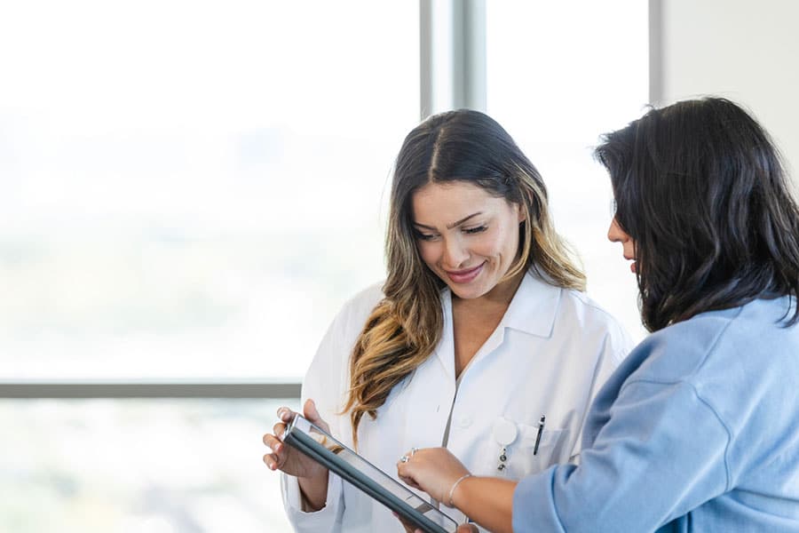 A doctor in a white coat smiles while looking at a tablet with a woman in a blue shirt, discussing Your Health together on the screen in a bright, modern office.