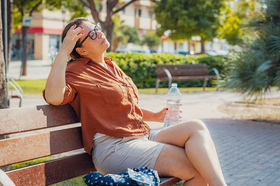 A woman sits on a park bench in the sun, holding a water bottle and shielding her face with her hand, possibly feeling hot or tired—a sign she could be experiencing heat exhaustion. Trees, benches, and a building are visible in the background.
