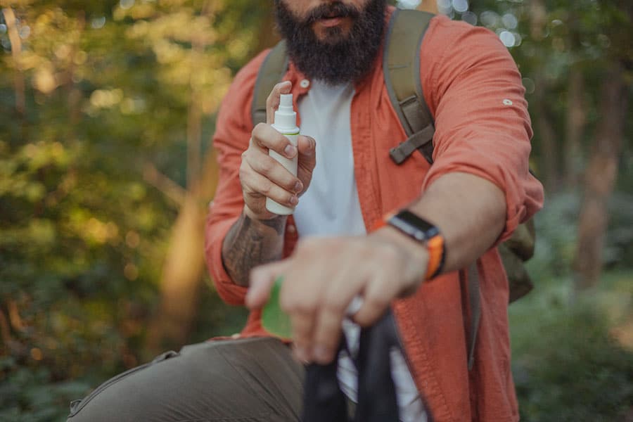 A bearded man wearing an orange shirt and backpack sprays insect repellent on his arm while outdoors in a forest, protecting himself from tick bites and the potential tick bite danger that can occur in wooded areas.