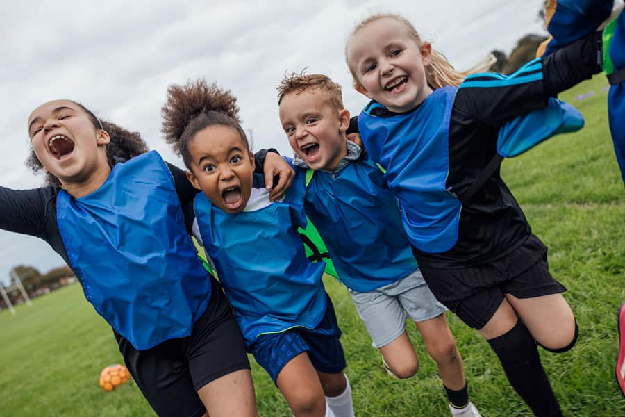 Four enthusiastic children in blue sports bibs smile and cheer with arms around each other, standing on green grass during a soccer game. A soccer ball and goal are visible in the background.