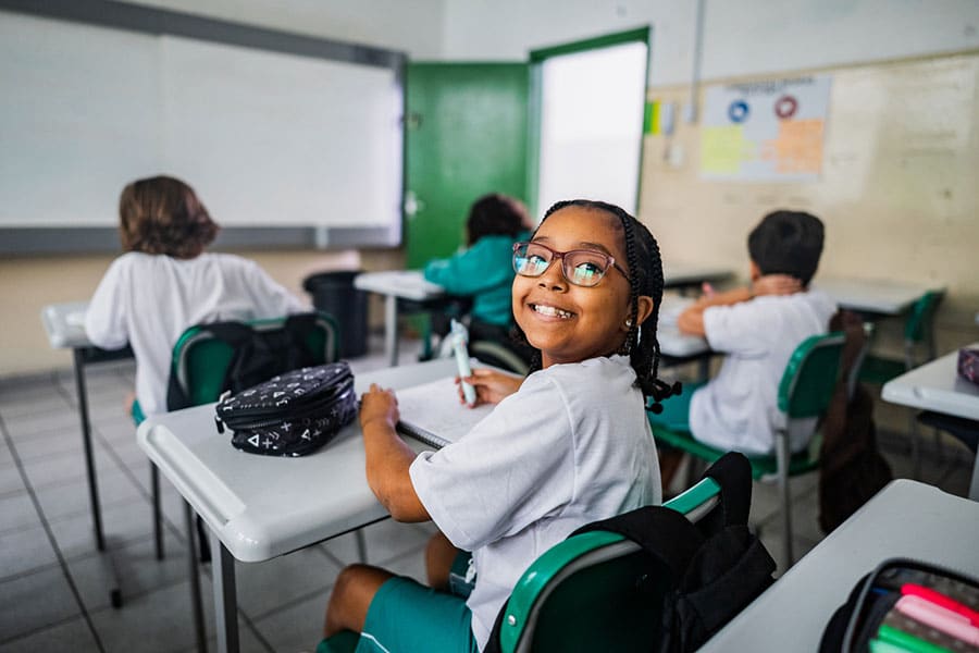 A smiling student with glasses sits at a desk in a classroom, looking at the camera, while other students work at their desks in the background—a perfect example of keeping healthy and engaged during the school year.