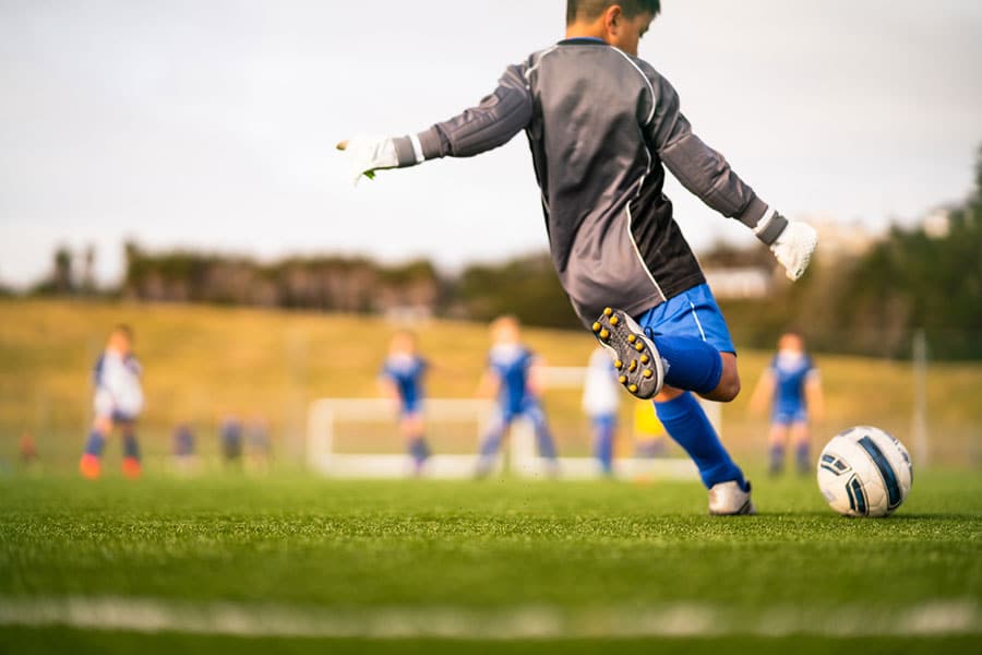 A youth soccer goalkeeper in gray and blue kicks a ball on a green field, representing the excitement of school sports, with teammates and a goal blurred in the background on a sunny day.
