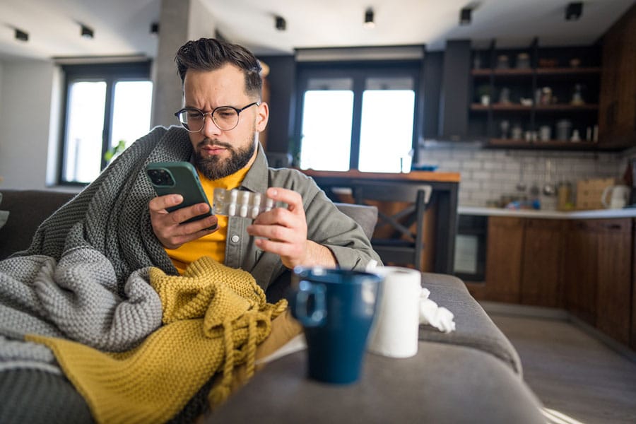 A man wrapped in blankets sits on a couch, looking at his phone while holding a glass of water. He appears unwell, with tissues and a mug on the table in a cozy, modern living room—clearly focused on treating a cold with home remedies.