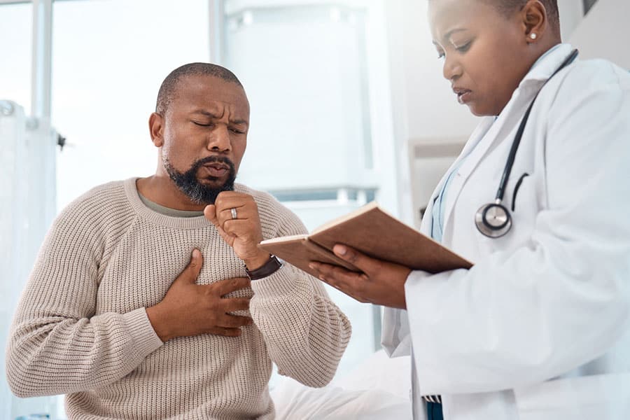 A patient coughing while holding his chest as a doctor reviews his medical records to assess pneumonia symptoms.